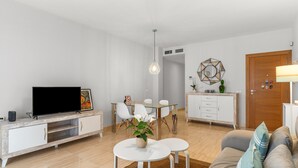 Een moderne woonkamer met een flatscreen televisie, witte meubels, een eettafel met stoelen, een bank en een decoratieve spiegel.