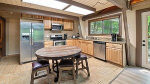 Una cocina con armarios de madera, electrodomésticos de acero inoxidable, una mesa redonda con taburetes y una ventana con vistas a árboles.