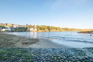 Una playa costera con un primer plano rocoso, una orilla arenosa y casas en una ladera al fondo.