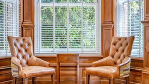 Deux fauteuils en cuir et une table en bois se trouvent dans une pièce dotée de grandes fenêtres et de murs lambrissés.