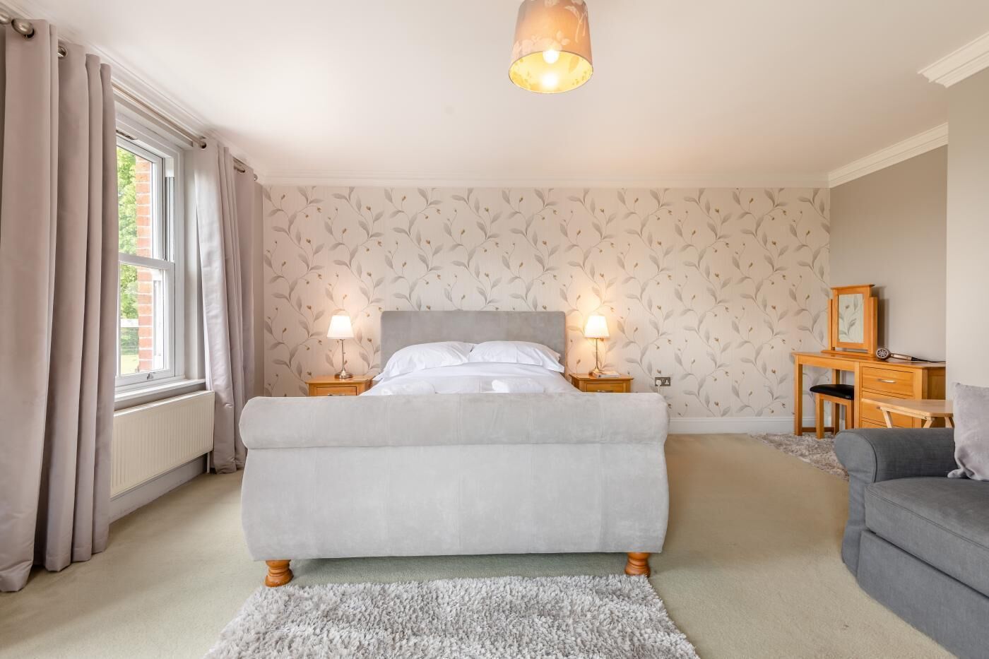 Une chambre à coucher avec un lit, deux tables de chevet avec des lampes, un canapé gris, un radiateur et une fenêtre avec des rideaux.
