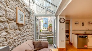 Una sala de estar acogedora con una pared de piedra, un sofá, una mesa de centro y vistas a una cocina y un espacio exterior a través de un ventanal.