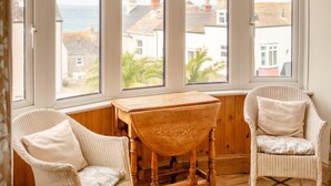 Una habitación con una mesa de madera y dos sillas de mimbre, un ventanal con vistas a casas y al mar.