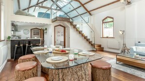Eine geräumige Küche mit hoher Decke und Oberlicht, ein Essbereich mit Glastisch und Hockern sowie eine Treppe, die zu einer oberen Ebene führt.
