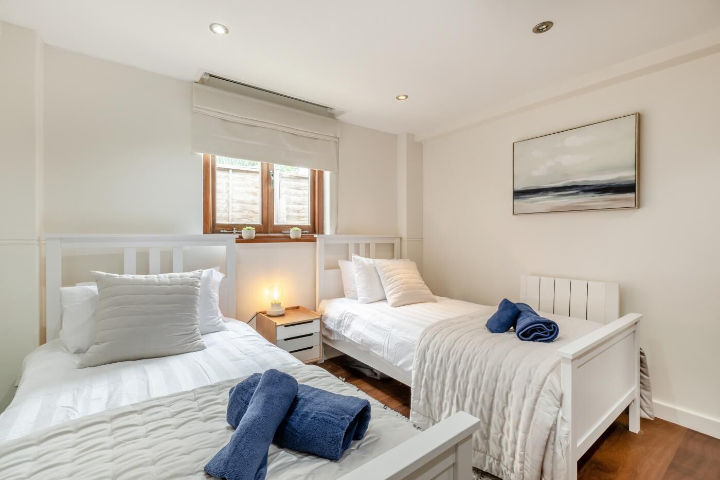 Quarto de hotel com duas camas, janela com cortinas, mesa de cabeceira com abajur e um quadro na parede.