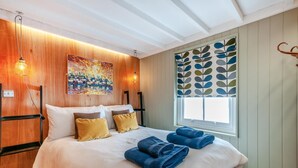 A hotel room with a wooden headboard, a large bed with white and yellow bedding, a colorful abstract painting, and a window with a leaf-patterned blind.