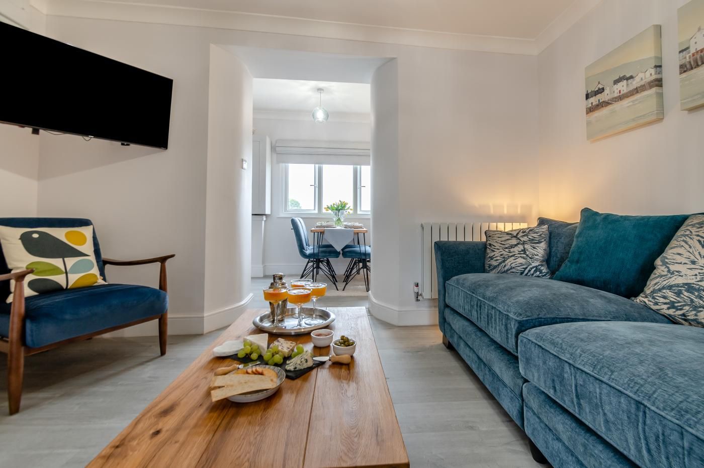 A modern living room with a blue sofa, a wooden coffee table with snacks, and a TV mounted on the wall.