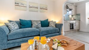 A living room with a sofa, a coffee table with food and drinks, and a kitchen in the background. - 2 Bed in Tenby (oc-c32240) (Tenby)