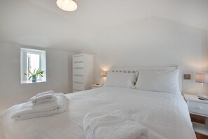 A neatly made bed with white linens and towels, a bedside table with a lamp, a tall white wardrobe, and a window with a plant. - 1 Bed in Porthleven (oc-crnest) (Porthleven)