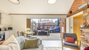 Una sala de estar moderna con un sofá, una mesa de comedor y una silla. Se observa una zona de cocina con un microondas y armarios. Se distingue una pared de ladrillo y una puerta de madera.
