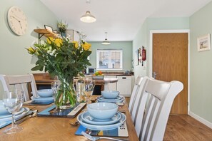 A dining area with a wooden table set for four, a vase with flowers, and a clock on the wall. - 2 Bed in Whitby (oc-w502) (Whitby)