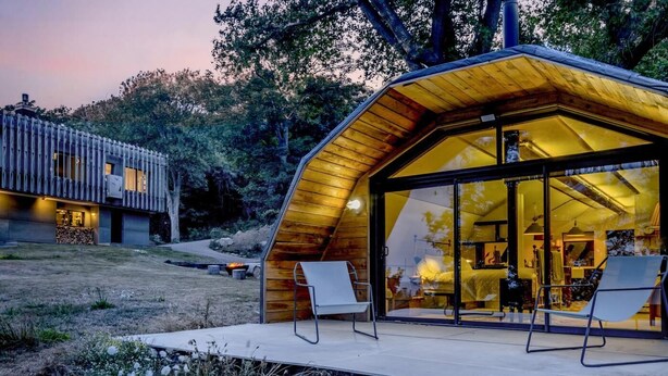 Uma cabana moderna com fachada de vidro, um deck com cadeiras e um braseiro no jardim.