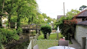 Vista junto al río con un puente de madera, muros de piedra y un jardín con una mesa y sillas.