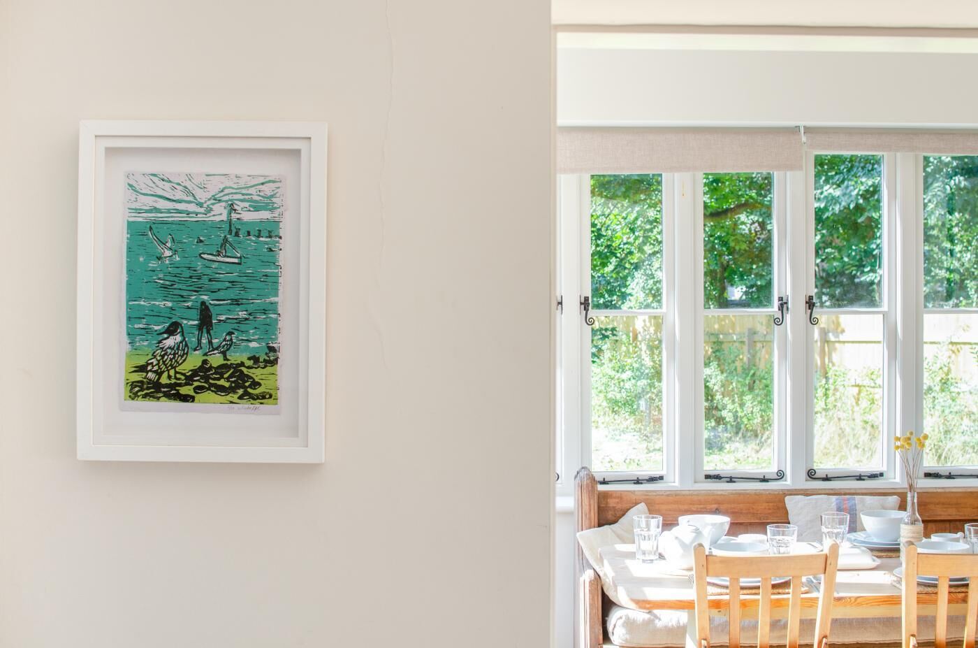 A dining area with a table set for two, a framed artwork on the wall, and a view through large windows.