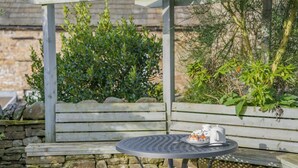 Una mesa metálica redonda con una tetera blanca y tazas, frente a una pared de piedra con bancos de madera.