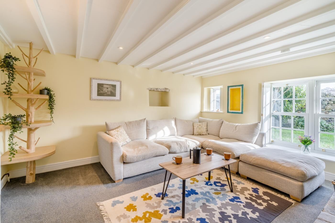 Ein gemütliches Wohnzimmer mit einem Ecksofa, einem Couchtisch, einem Holzregal für Pflanzen und einem großen Fenster mit Blick auf Grünflächen.