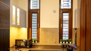 A compact kitchen with a microwave, sink, and cabinets. There are two windows with wooden frames and a small potted plant on the windowsill.