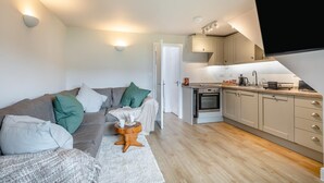 A compact living space with a sofa, a small round table with a cup, a kitchen area with a stove and cabinets, and a TV mounted on the wall. - 3 Bed in Corton (oc-s32347) (Corton)