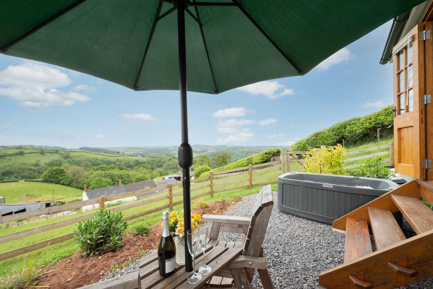 A patio with a hot tub, a wooden table with a bottle and glasses, and a view of rolling hills.