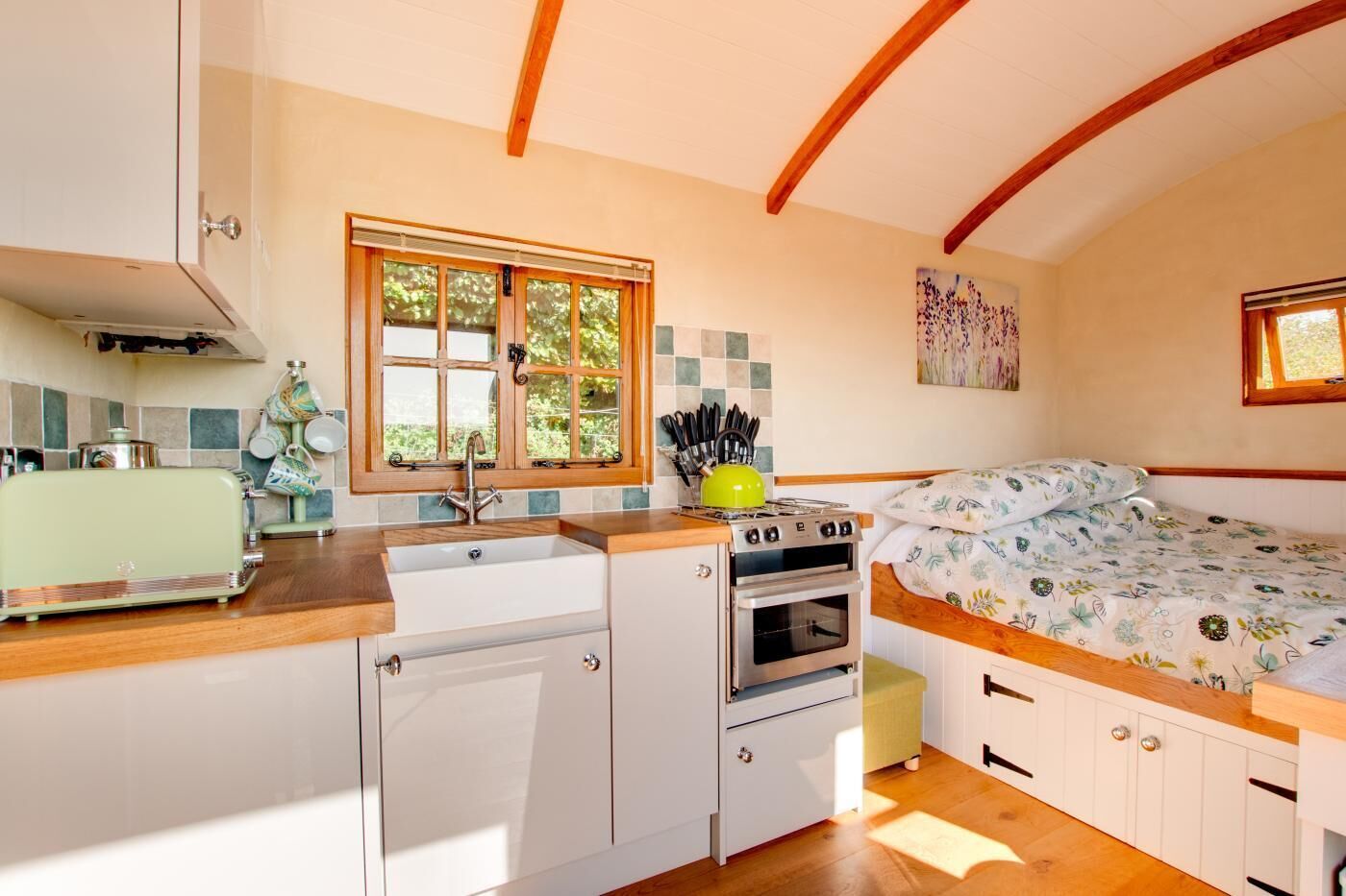 A compact kitchen with a window, a sink, a stove, and a bed with floral bedding.