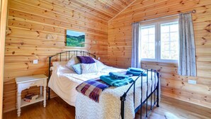 A wooden cabin bedroom with a bed, a nightstand, a window with curtains, and a painting on the wall. - 1 Bed in Pickering (oc-y290) (Middleton)