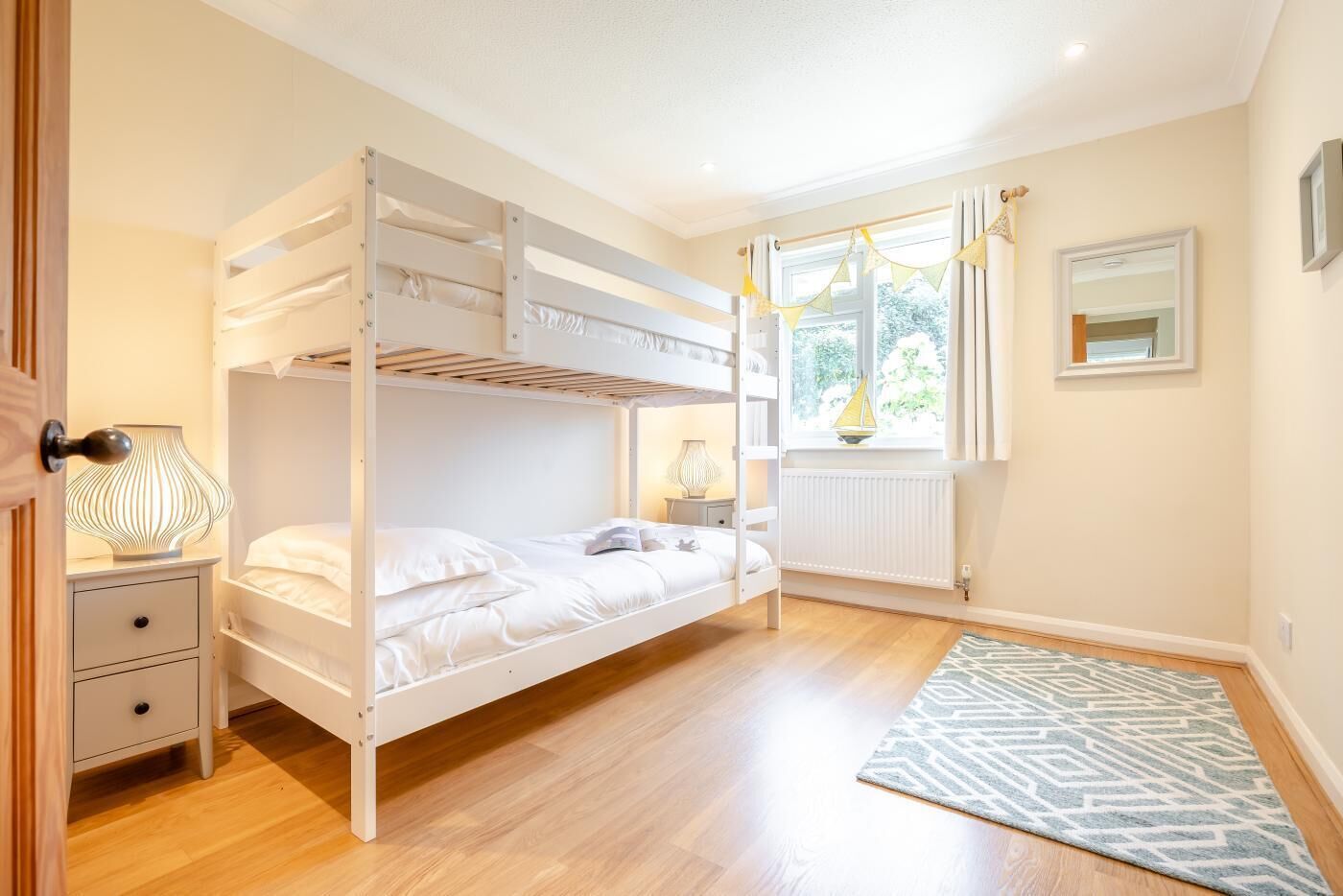 A bunk bed room with a wooden floor, a bedside table with a lamp, a window with curtains, and a radiator.