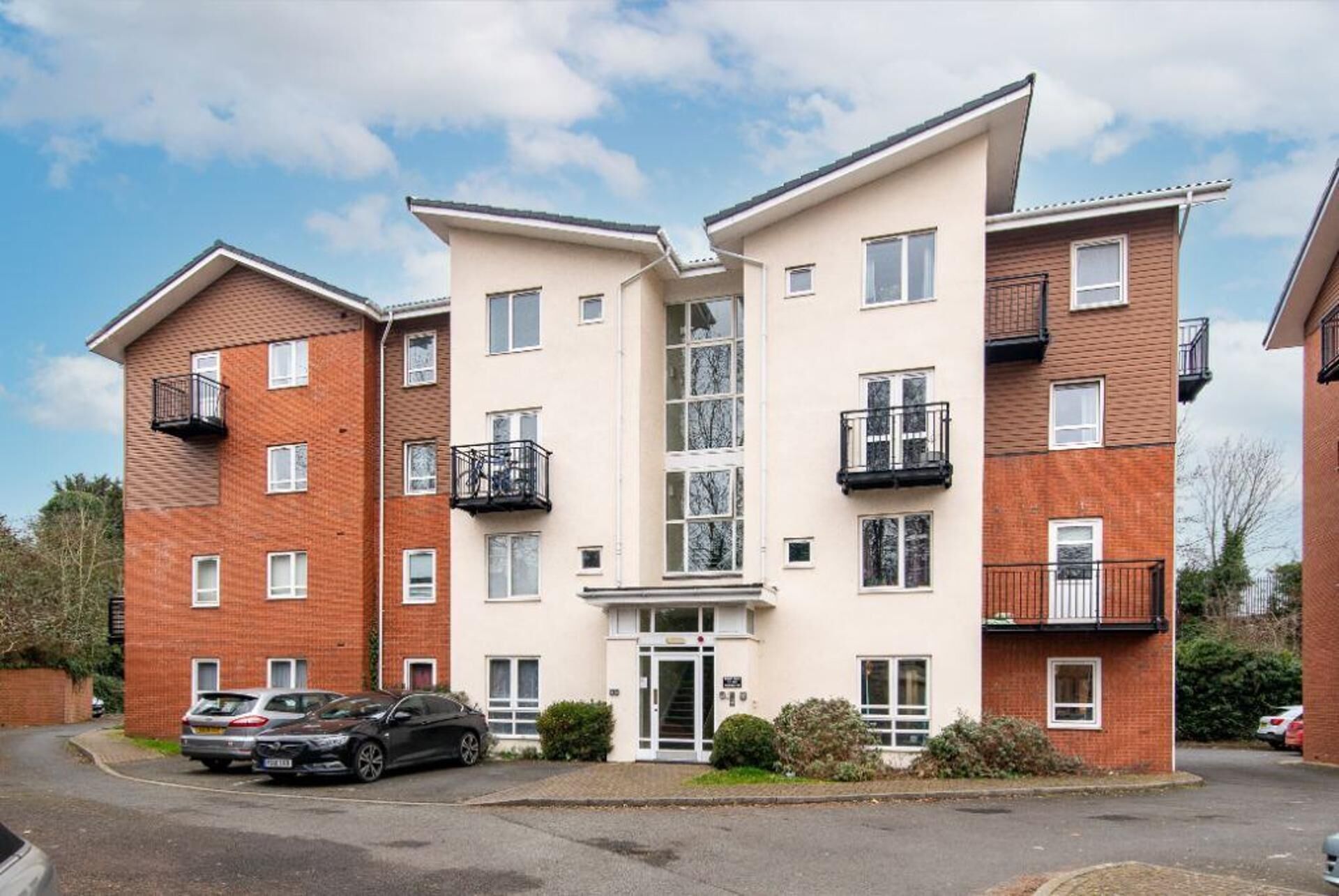 A multi-story residential building with a central entrance, balconies, and a parking area with cars.