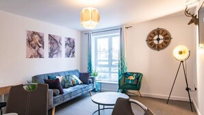 A modern living room with a sofa, a round coffee table, and a chair. There are abstract paintings on the wall, a clock, and a floor lamp. - Apartment - Sleeps 2 - Parking - City Centre Close (Coventry)
