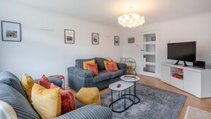 Un salon moderne avec un canapé gris, des coussins colorés, une petite table ronde, un téléviseur posé sur un meuble blanc et des tableaux encadrés accrochés au mur.