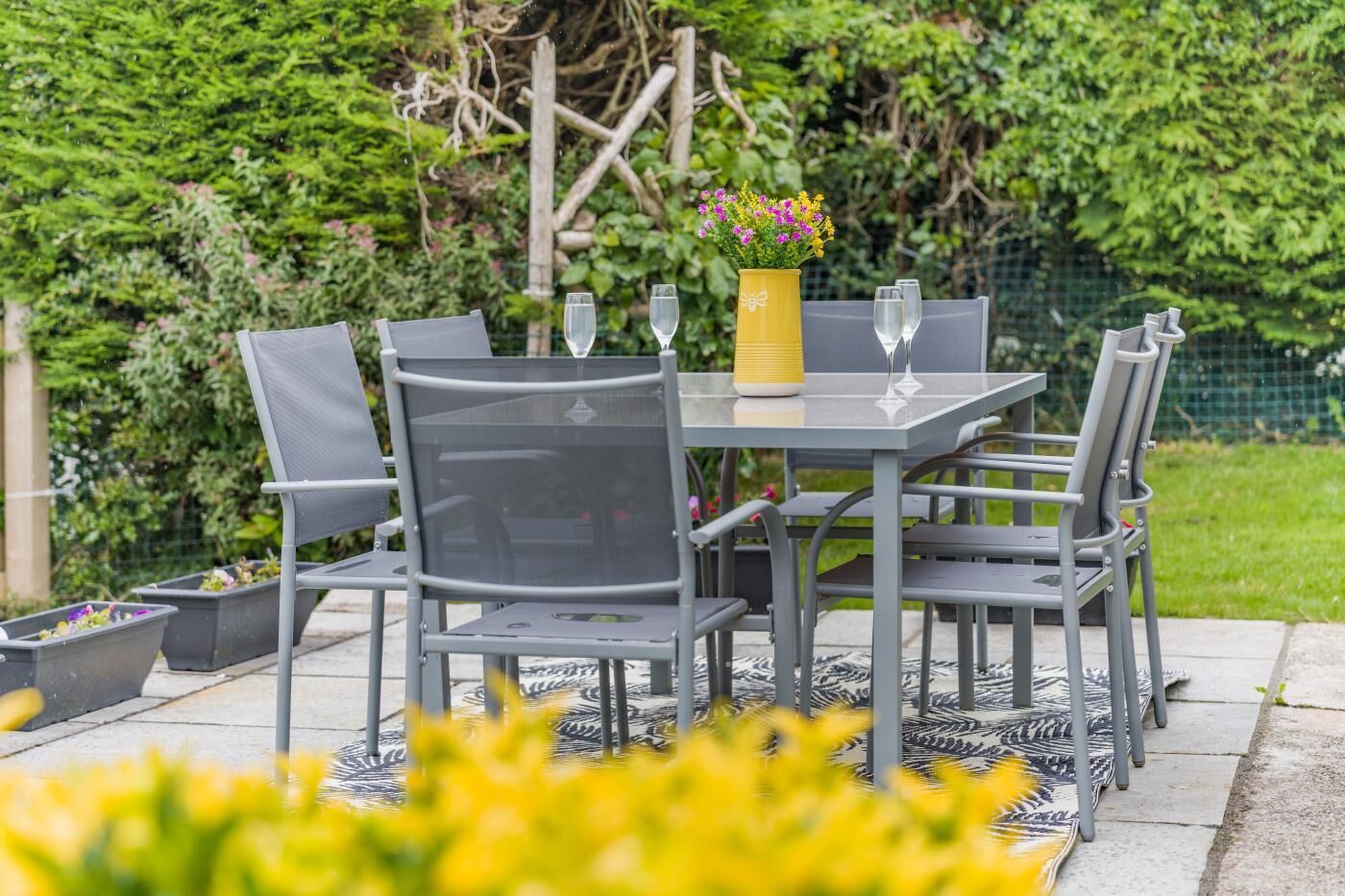An outdoor patio with a grey metal table and chairs, a yellow vase with flowers, and a glass of water on the table.