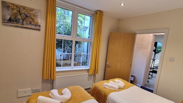 A hotel room with two beds, each with a white comforter and a rolled towel. There is a window with yellow curtains, a radiator below, and a painting on the wall. - Coventry Retreat - Modern 2 Bed Apartment (Coventry)