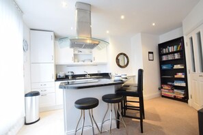 A modern kitchen with a central island, bar stools, a bookshelf, and a clock on the wall. - 2 Bed in Whitby (oc-w026) (Whitby)