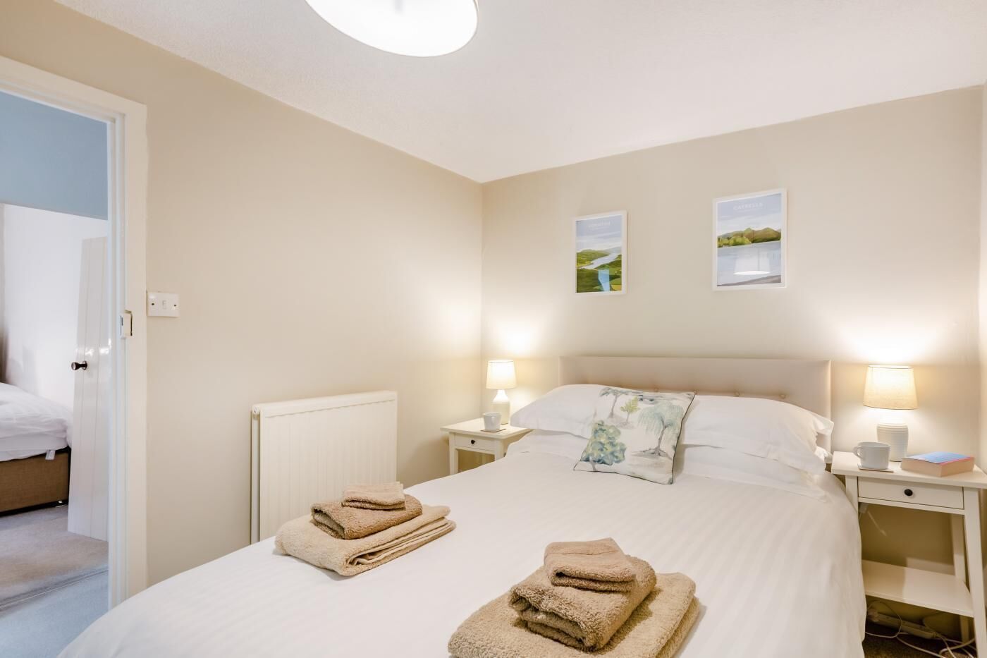A neatly made bed with two pillows, a bedside table with a lamp, and a wall with two framed pictures.