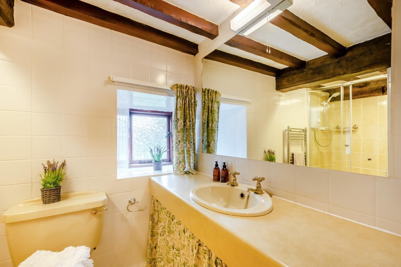 A bathroom with a toilet, sink, and shower. There is a window with curtains, a mirror, and a plant on the counter.