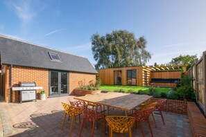 A backyard with a wooden deck, a barbecue grill, and a dining area with chairs. - 5 Bed in Southwold (oc-nort) (Southwold)