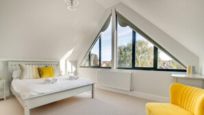 Ein Schlafzimmer mit schrägen Wänden, einem Bett mit weißer Bettwäsche, einem gelben Sessel und einem Fenster mit Blick auf Bäume.