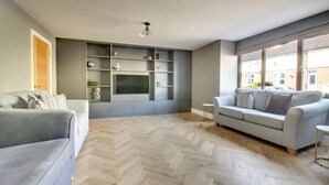 Ein modernes Wohnzimmer mit einem grauen Sofa, einem an einer grauen Wand befestigten Fernseher und einem Holzboden mit Fischgrätmuster.