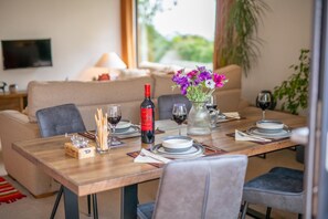 Ein Esszimmer mit Holztisch, gedeckt für vier Personen, einer Weinflasche, einer Blumenvase und einer Couch im Hintergrund.