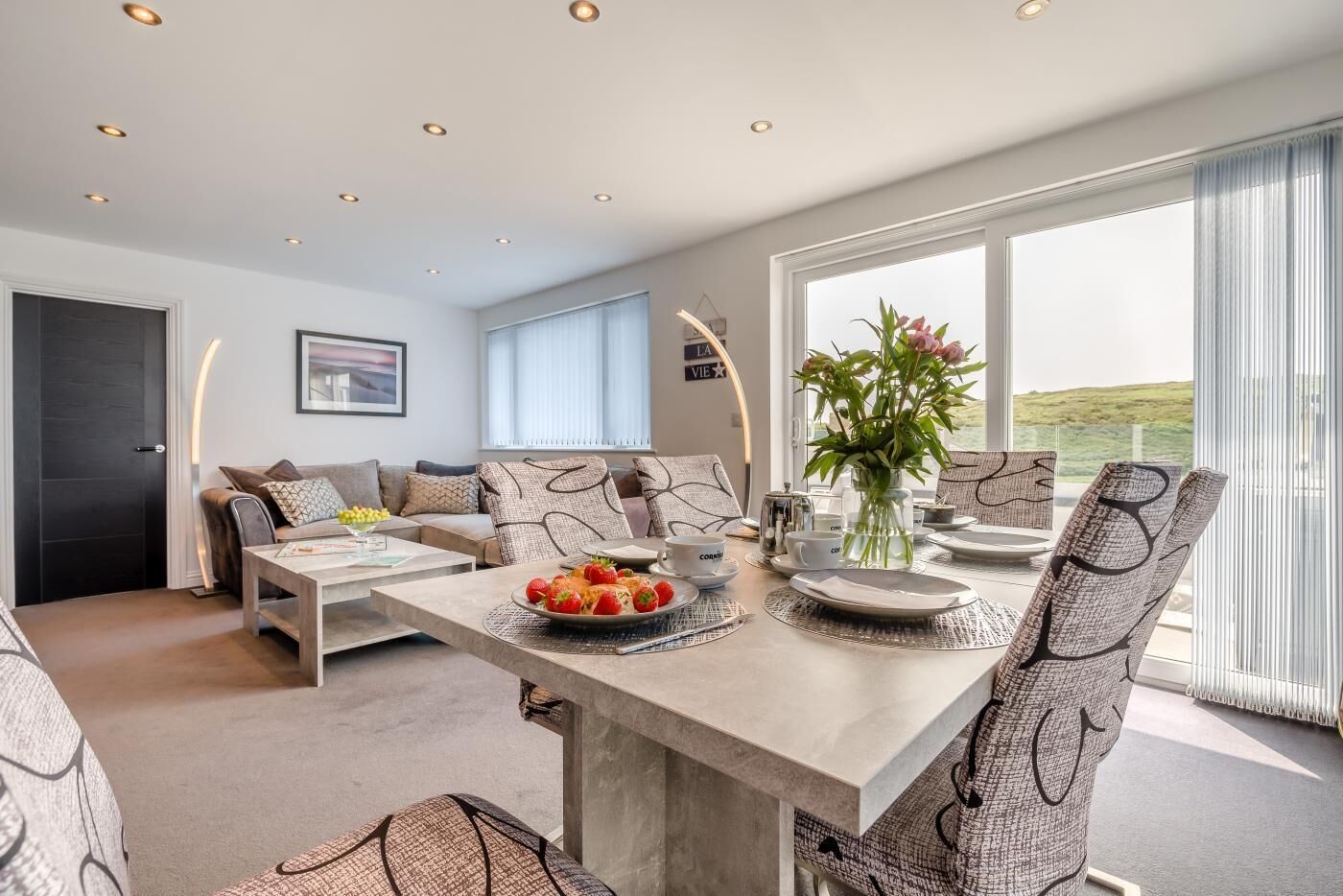 A modern living room with a dining area, featuring a sofa set, a coffee table with fruit, and a view of the outdoors.
