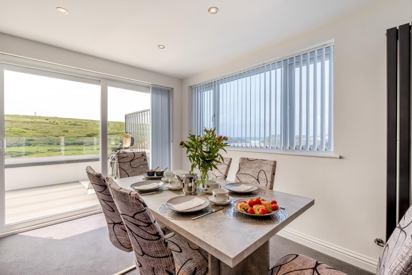A dining area with a table set for four, large windows, and a view of a green landscape.