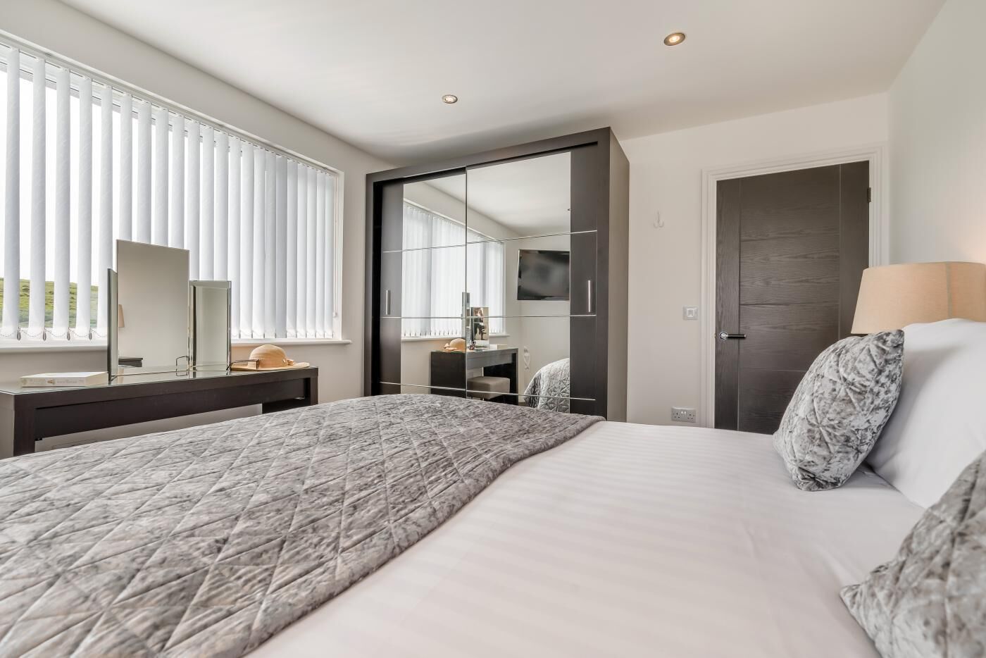 A modern hotel room with a large bed, a desk with a mirror, a TV, and a wardrobe.