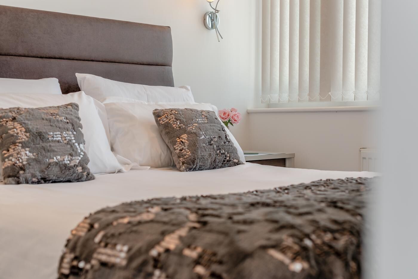 A neatly made bed with white linens and decorative pillows, a headboard, a bedside table, and a window with vertical blinds.