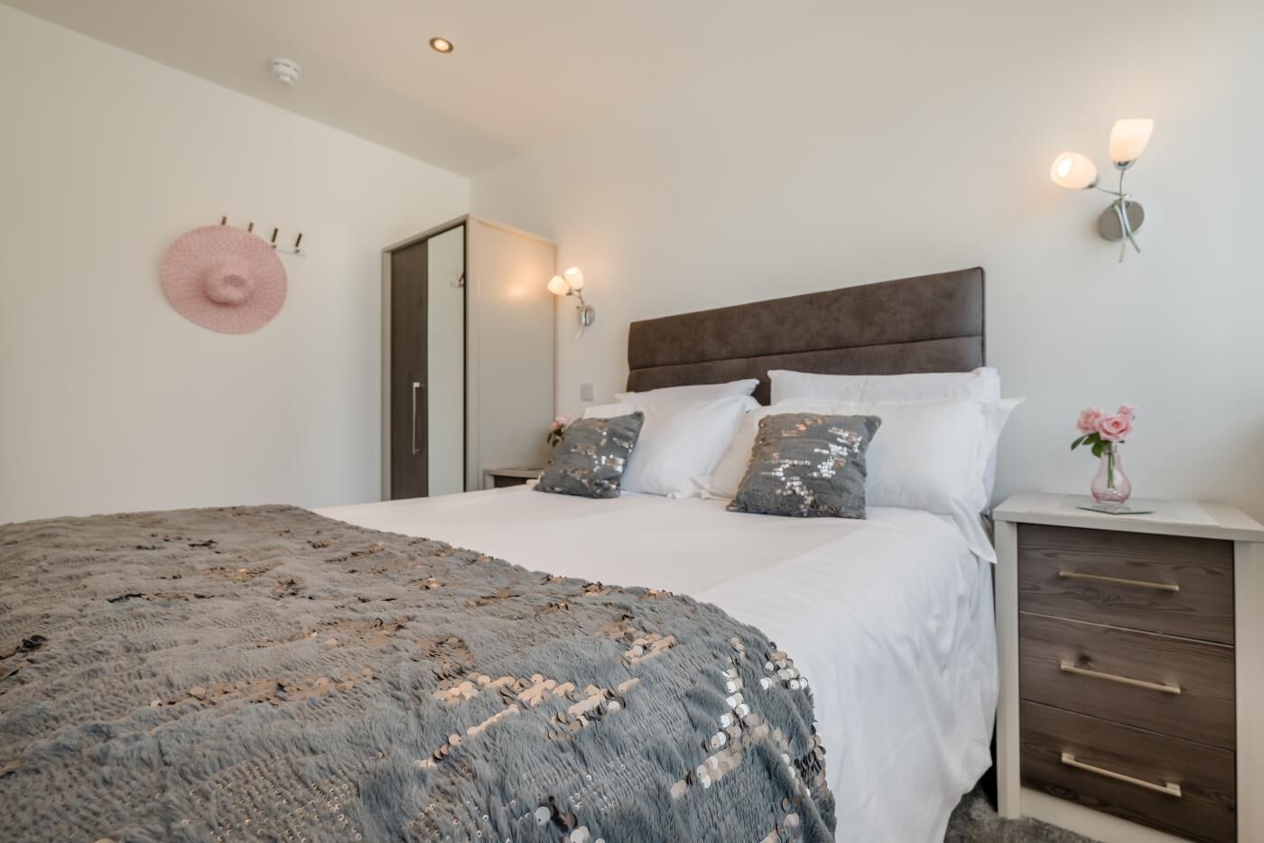 Een slaapkamer met een groot bed, een nachtkastje met een bloevas en een wandlamp.