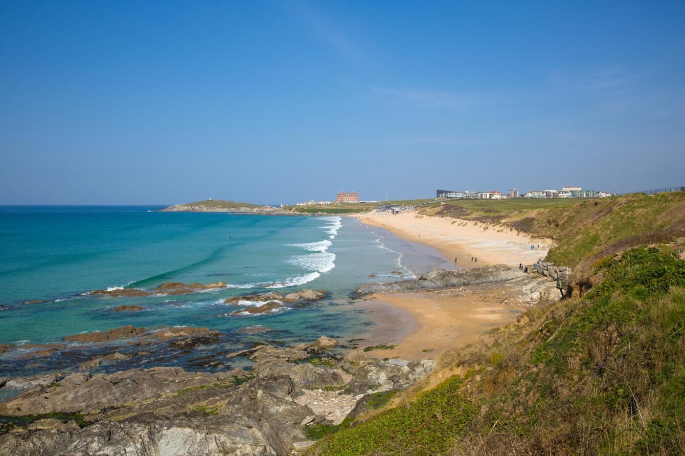 Ein Küstenstrand mit felsigem Ufer, klarem, blauem Wasser und Gebäuden im Hintergrund.