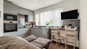 A compact kitchen with modern appliances, a dining area with a small table and chairs, and a large window with blinds. - 1 Bed in Newquay (oc-o32401) (Holywell)