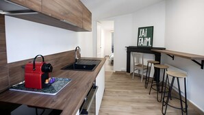 A modern kitchen with a wooden countertop, a red kettle, and a sink. There are two stools and a small table with a sign that reads "J'AI FAIM." - Le perchoir de Maurice, calme et cosy (Arras)