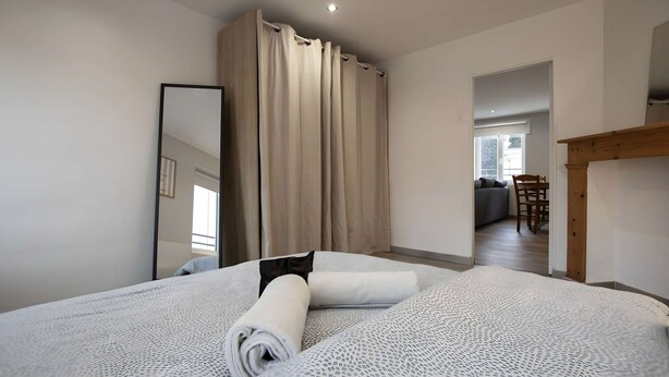 A bedroom with a large bed, a wooden headboard, a mirror, and a view into a living area with a sofa and a dining table. - Le perchoir de Maurice, calme et cosy (Arras)