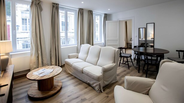 Ein modernes Wohnzimmer mit einer Couch, einem Holztisch, einem Esstisch mit Stühlen und großen Fenstern mit Vorhängen.