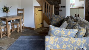 Ein gemütliches Wohnzimmer mit einer Holztreppe, einer Steinwand, einem Essbereich mit Holztisch und -stühlen sowie einem gemusterten Sofa.
