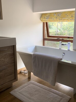 Une salle de bain avec une baignoire sur pieds, un meuble-lavabo en bois et une fenêtre agrémentée d’un store à motifs floraux jaunes.